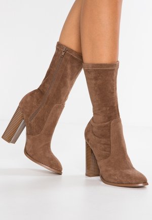 Braune Wildleder-Stiefel bis zur Mitte der Wade mit Blockabsätzen und seitlichen Reißverschlüssen, getragen an übereinandergeschlagenen Beinen vor weißem Hintergrund.