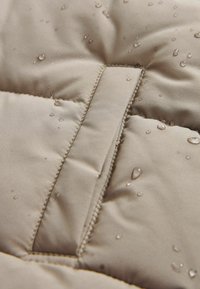Gros plan sur un tissu matelassé beige avec des gouttes d'eau à la surface, mettant en avant les coutures déperlantes et la texture.