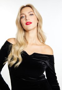 Sort fløjl af skuldrene kjole med V-formet udskæring, lange ærmer og tætsiddende silhuet. Modellen har bølget blond hår og rød læbestift.