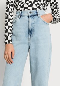 Jeans de ganga de cintura alta azul claro com corte reto, apresentando um design clássico de cinco bolsos e um acabamento desbotado, usados com uma blusa de manga longa estampada.
