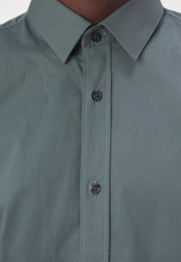 Camicia grigia con bottoni e colletto, caratterizzata da bottoni scuri e una texture del tessuto liscia. Cuciture visibili lungo la patta dei bottoni.