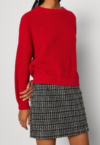 Maglione rosso a maglia con scollo ampio e vestibilità comoda, abbinato a una gonna in tweed nero e bianco a trama. 