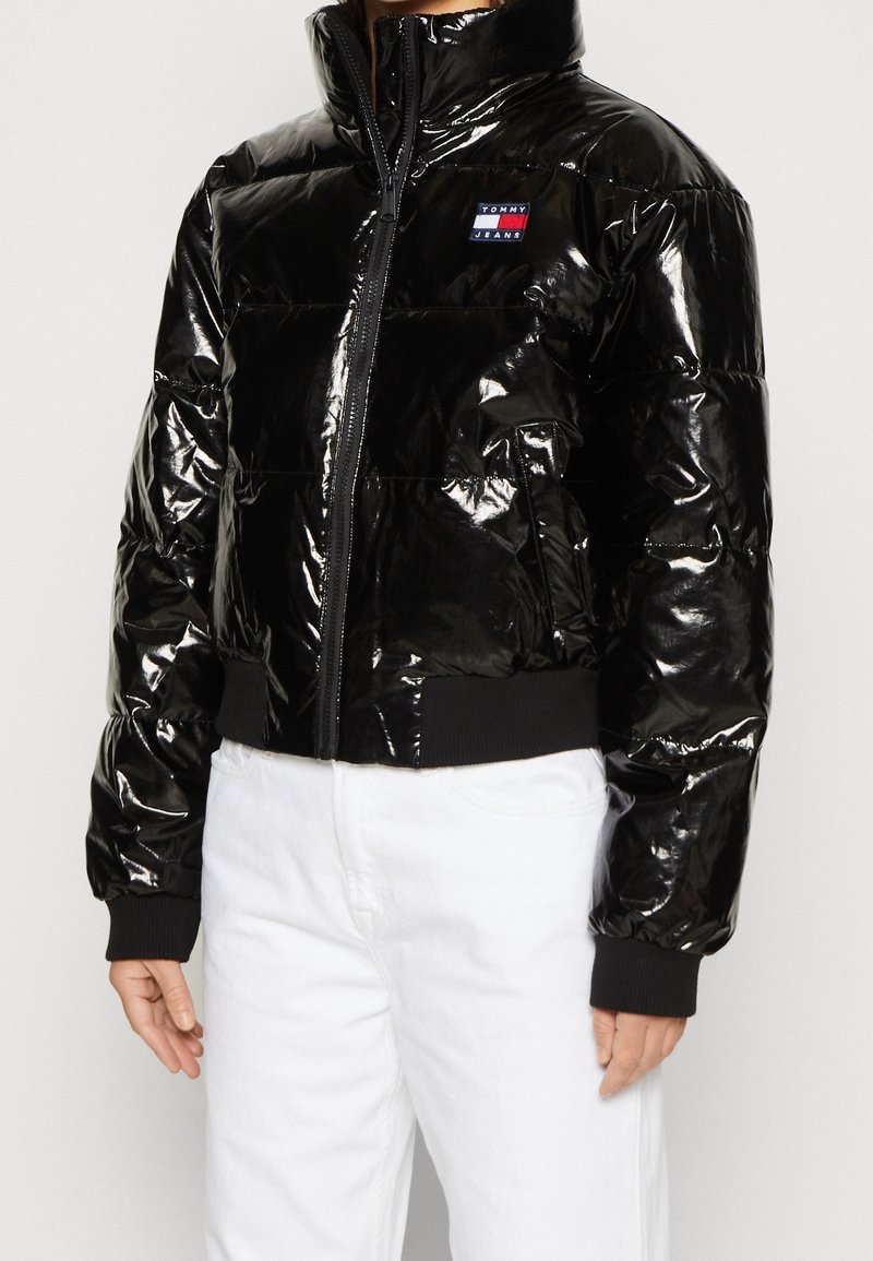Veste matelassée noire brillante avec fermeture éclair frontale et logo "Tommy Jeans", portée sur un pantalon blanc, montrant partiellement le torse et les bras.