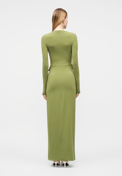 Γυναίκα με μακριά ίσια μαλλιά που φοράει εφαρμοστό μακρύ φόρεμα σε ελιά πράσινο με μακριά μανίκια και λευκά παπούτσια με ψηλό τακούνι, βλέπουμε από πίσω.