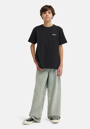 Junger Junge steht und trägt ein schwarzes T-Shirt mit Grafik, hellblaue Schlagjeans und schwarze Low-Top-Sneaker vor weißem Hintergrund.