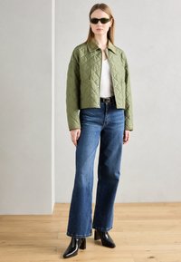 Jachetă de culoare verde măslinie, căptușită, tricou alb, blugi evazați albaștri și cizme negre cu toc. Accesoriile includ ochelari de soare eleganți, de culoare închisă.