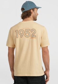 Lys gul kortærmet t-shirt med et stort "1952" grafisk tryk i orange og sort på ryggen, lavet af blødt bomuldsstof.