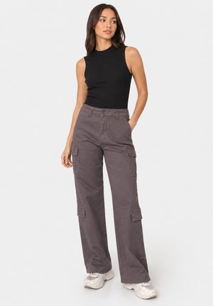 Donna che indossa un top nero senza maniche e pantaloni cargo grigi con tasche, in piedi con una mano in tasca e guardando di lato.