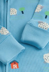Tela azul acanalada con ovejas, árboles verdes y un granero rojo estampados en puños y mangas suaves de ropa para bebés.