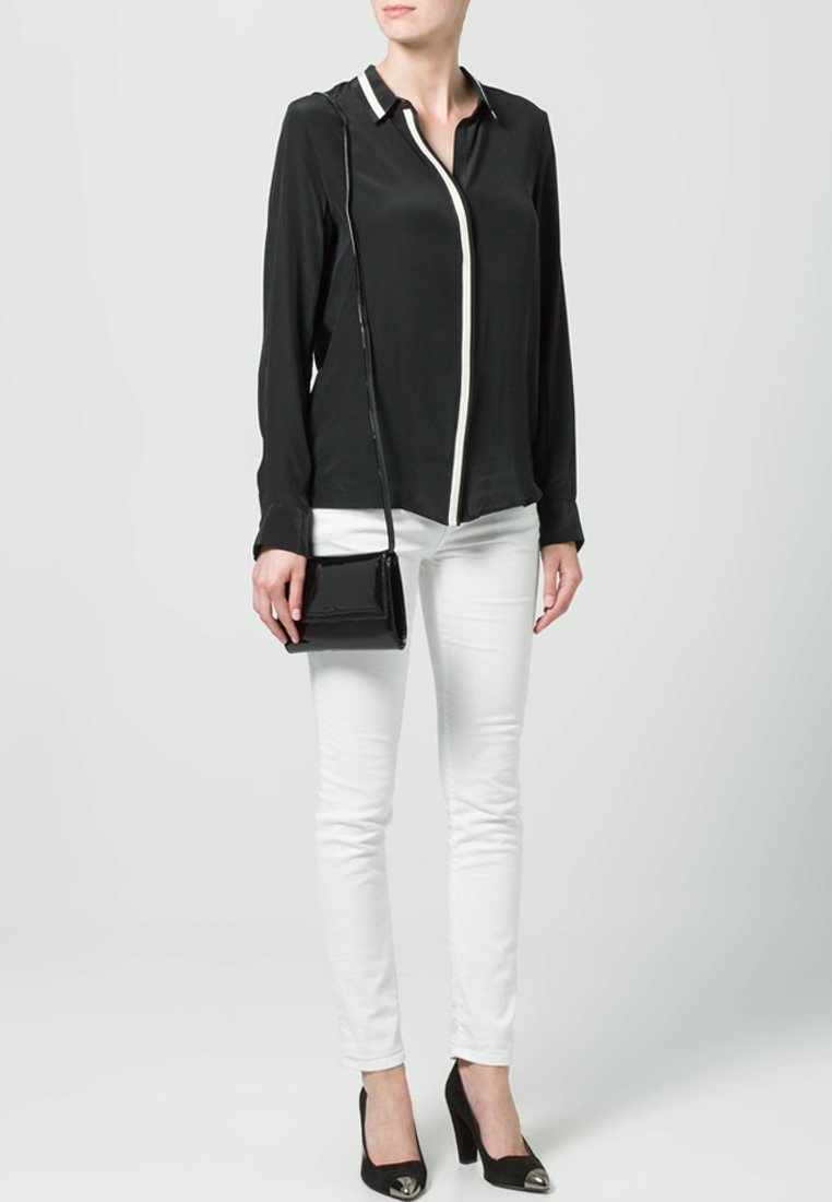 Mujer vestida con blusa negra de manga larga con ribete blanco, pantalones blancos ajustados, tacones negros con puntas metálicas y un bolso negro de hombro.
