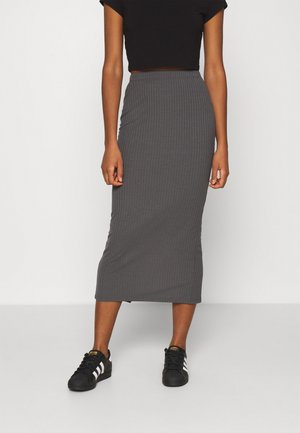 Person trägt einen grauen, gerippten, hoch taillierten Midirock mit einem schwarzen Crop-Top und schwarzen Adidas-Sneakern mit weißen Streifen.