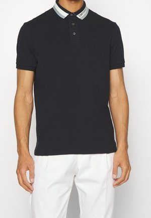 Mann trägt ein schwarzes Poloshirt mit kurzen Ärmeln, gestreiftem Kragen und dem Schriftzug "EMPORIO", kombiniert mit weißen Hosen.