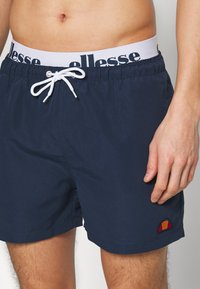 Maritime svømmebukser laget av lett materiale, med en hvit snor og elastisk linning med svart skrift, pluss en fargerik logo.