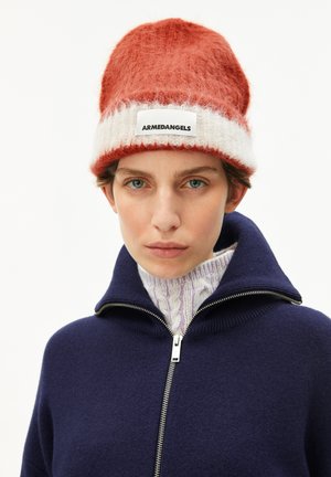 Une personne avec les cheveux courts porte un bonnet en tricot rouge et blanc et un pull zippé bleu marine sur un col roulé en tricot torsadé violet clair.