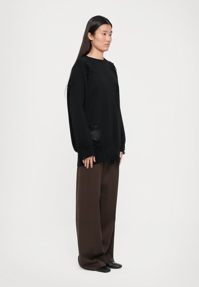 Black oversized sweater with frayed accents and mesh pockets, paired with wide brown trousers and black shoes, on a plain background.