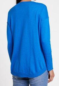 Blauwe gebreide trui met een ronde halslijn, afgehaakte schouders en geribbelde manchetten, met een ontspannen pasvorm. Gdragen met lichtgewassen jeans.