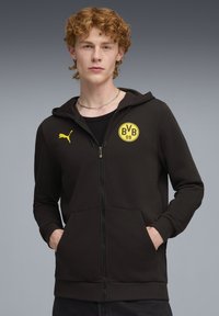 Sort zip-up hættejakke lavet af blødt stof, har en kængurulomme, ribbede manchetter og et gult rundt logo med "BVB 09" på brystet.