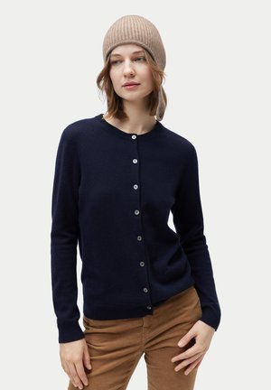 Frau, die eine marineblaue, geknöpfte Strickjacke, braune Hose und eine beige gestrickte Mütze vor einem schlichten hellen Hintergrund trägt.