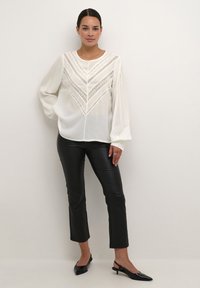 Blouse blanche avec un motif en chevrons texturé et des accents en dentelle, associée à un pantalon en cuir noir raccourci et des escarpins noirs à talons aiguille.