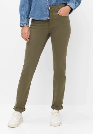 Personne portant un pantalon droit vert olive retroussé aux chevilles, des baskets blanches et une chemise en jean bleue avec une main dans la poche.