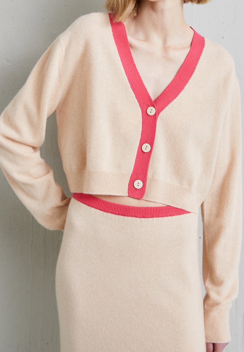 Vaaleanbeige lyhyt cardigan, jossa on syvä V-pääntie, kontrastina vaaleanpunaiset reunat ja puunapit. Pehmeä rakenne, yhdistettynä samaan hameeseen.
