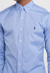 Muž v světle modré košili na knoflíky s tmavě modrým logem póla na hrudi, s knoflíkem u límce zapnutým.