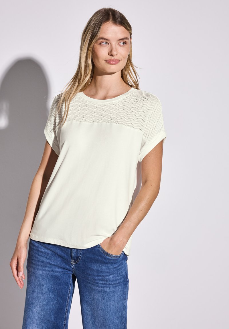 Lys creme farvet kortærmet top med struktureret strik yoke og glat krop. Parret med blå jeans, står med hænderne i lommerne.