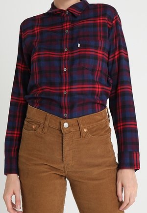 Personne portant une chemise à carreaux rouge et marine boutonnée, rentrée dans un pantalon en velours côtelé marron taille haute, les mains détendues le long du corps.