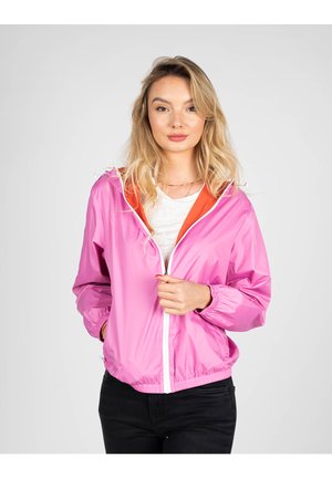 Giacca a vento fucsia con zip bianca e polsini elasticizzati, dotata di cappuccio foderato arancione. Indossata sopra una maglietta bianca, abbinata a jeans neri.