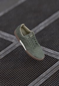 Sapatilha verde de camurça com um design de atacadores, sola de borracha castanha e meio-sola branca. Apresenta três riscas de lado e um superior texturizado.