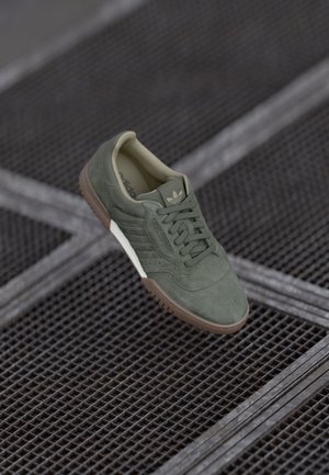 Roheline sametist sneaker, millel on nööpidega disain, pruun kummist tald ja valge vahepealne tald. Külgedel on kolm riba ja tekstuuritud ülemine osa.