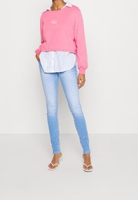 Mujer vestida con jeans ajustados de un azul claro, tacones abiertos blancos, sudadera rosa en capas sobre una camisa de botones a rayas, manos en los bolsillos.