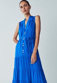 Sininen hihaton mekko, jossa on nappilista edessä, vyötärölle sidottava solmio ja kerrostettu muotoilu. Kangas on ruudukkomuotoiltu, siinä on sileä pinta ja pehmeä laskeutuminen.