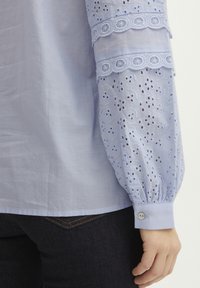 Svetlo modra bluzica z vezenimi detajli v očesni zasnovi na rokavih, napihnjene oblike in zapenjanjem na manšeti. Gladek, zračen material.