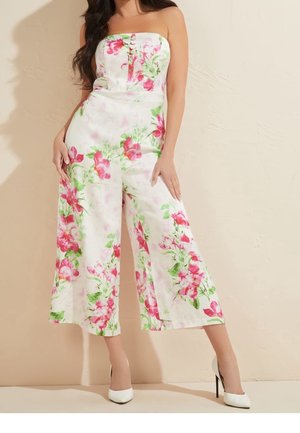 Frau trägt weißen trägerlosen Jumpsuit mit rosa-grünem Blumenmuster, kombiniert mit weißen High Heels, steht vor einer schlichten Wand.