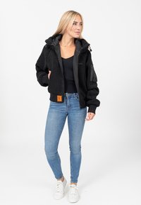 Veste zippée en polaire noire avec une surface texturée, dotée d'une capuche, de poignets élastiques et d'une poche latérale. Portée par-dessus un haut noir ajusté et un jean.