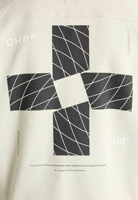 Világosszürke pulóver nagy fekete geometriai mintával, fehér díszítésekkel, szöveges elemekkel és textúrált mintázattal.