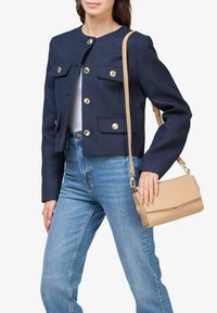 Chaqueta corta azul marino con botones dorados y dos bolsillos frontales, combinada con jeans de cintura alta en azul claro y un bolso cruzado beige.