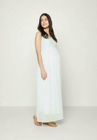 Vestido de maternidade verde claro com um design fluido e decote em V, apresentando uma saia longa e um tecido suave. Combinado com sandálias castanhas.