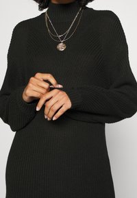 Svart stickad tröja med hög hals, ribbat mönster och l ånga ärmar. Har lager av guldkedjor och släta, välvårdade händer.