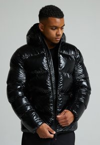 Giacca a vento nera con un esterno lucido e trapuntato, cappuccio e zip frontale. Presenta un design attillato e polsini elasticizzati.