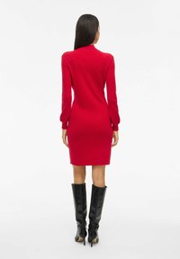 Robe en tricot rouge avec un col haut, des manches longues et une silhouette ajustée, associée à des bottes noires à talons hautes arrivant aux genoux. Texture lisse, motif côtelé.