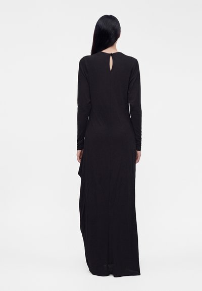 Femme aux longs cheveux noirs portant une longue robe noire à manches longues, vue de dos sur un fond blanc uni.