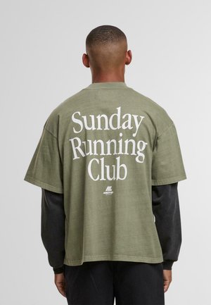 Mies, jolla on lyhyt hius, pukeutunut oliivinvihreään t-paitaan mustan pitkähihaisen paidan päällä, selässä teksti "Sunday Running Club."