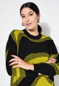 Maglione nero con un audace motivo giallo a onde. Tessuto a maglia, scollatura a girocollo e vestibilità rilassata. Modello che mostra anelli e smalto nero sulle unghie.