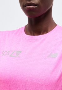 Κοντινό πλάνο ενός ατόμου που φοράει ροζ αθλητική μπλούζα με διάτρητο σχέδιο και γκρι λογότυπα "tcs LZT" και New Balance στο στήθος.