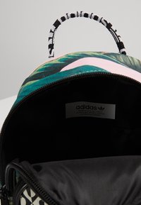 Zielono-różowy plecak z tropikalnym wzorem liści, czarnym wnętrzem i wzorzystym uchwytem. Komora na zamek błyskawiczny odsłania metkę marki.