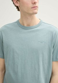 Tricou bluet deschis cu guler rotund, realizat dintr-un material moale și texturat; dispune de un detaliu mic, embosat cu logo pe piept. Mâneci scurte.