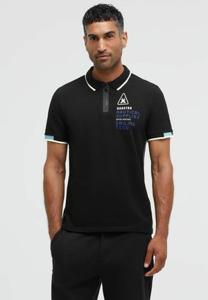 Man wearing black polo shirt with white trim and nautical-themed text on chest, standing with one hand in pocket against gray background.