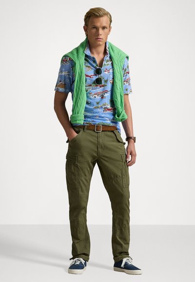 Herenoutfit met een blauwe gestreepte shirt, een groene trui over de schouders, olijfgroene cargobroek en blauwe sneakers met witte zolen.